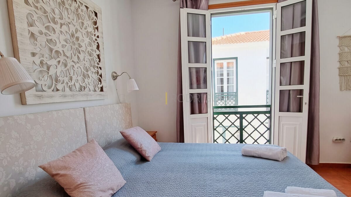 Helles Schlafzimmer mit dekorativem Kopfteil, großen Fenstern mit Balkonzugang und Makramee-Wandkunst.