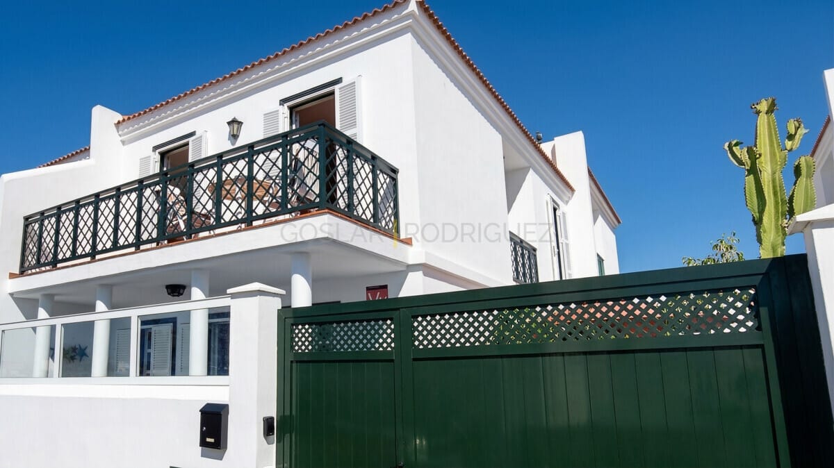 Moderne weiße Villa mit Balkon, grünes Tor und Kaktus, sonniger Tag.
