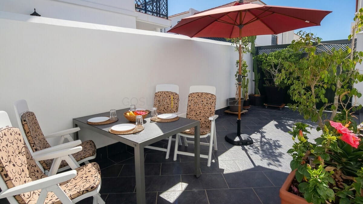 Essbereich im Freien mit Tisch, Stühlen und Sonnenschirm. Tropische Pflanzen und sonnige Atmosphäre.