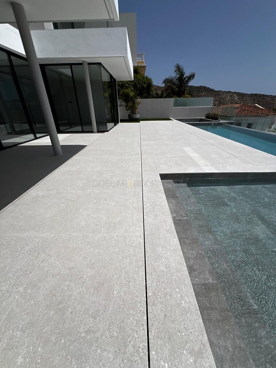 Aussenaufnahme einer modernen Villa in Adeje, Teneriffa, mit Pool. Die Aufnahme zeigt die Terrasse mit hellgrauen Fliesen, den Pool und einen Teil des Hauses mit grossen Glasfronten sowie einen Teil der Landschaft.