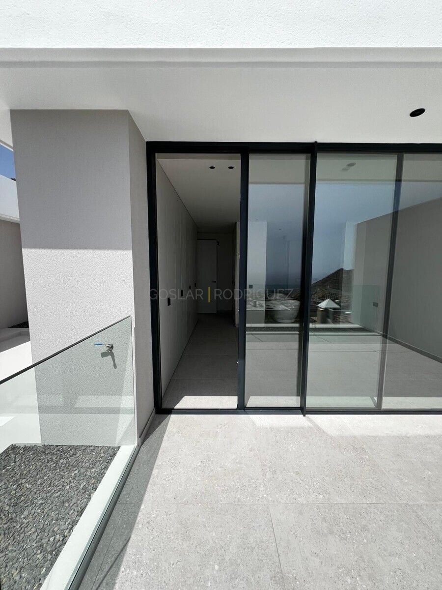 Blick auf eine moderne Villa in Adeje auf Teneriffa. Das Bild zeigt eine offene Terrasse mit Glasgeländer und Schiebetüren, die einen hellen Innenraum mit Einbauschränken freigeben. Die Villa bietet einen Ausblick.